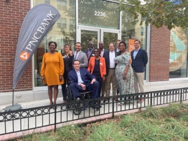 A group of people stands outside a building adorned with a PNC sign. One person is in a wheelchair. Attire varies from formal to business casual, adding depth to the scene. Trees and another building provide a serene backdrop.